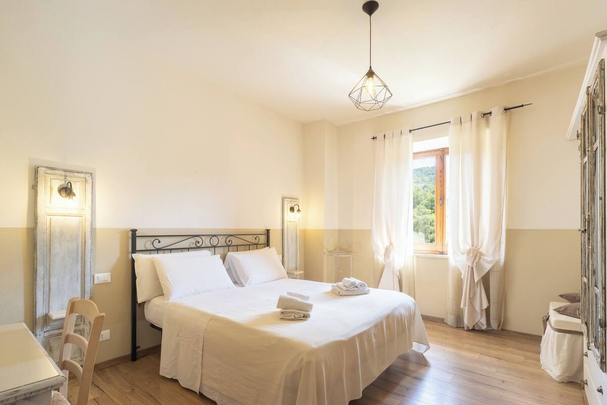 Une chambre bien rangée, avec un lit, des tables de chevet, une fenêtre avec des rideaux et un luminaire suspendu.