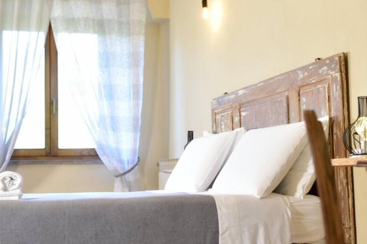 Un lit bien fait, avec du linge de lit blanc, une tête de lit en bois et une fenêtre ornée de rideaux légers.