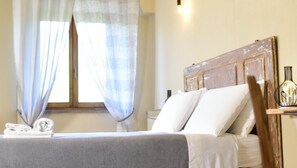 Un lit bien fait, avec du linge de lit blanc, une tête de lit en bois et une fenêtre ornée de rideaux légers.