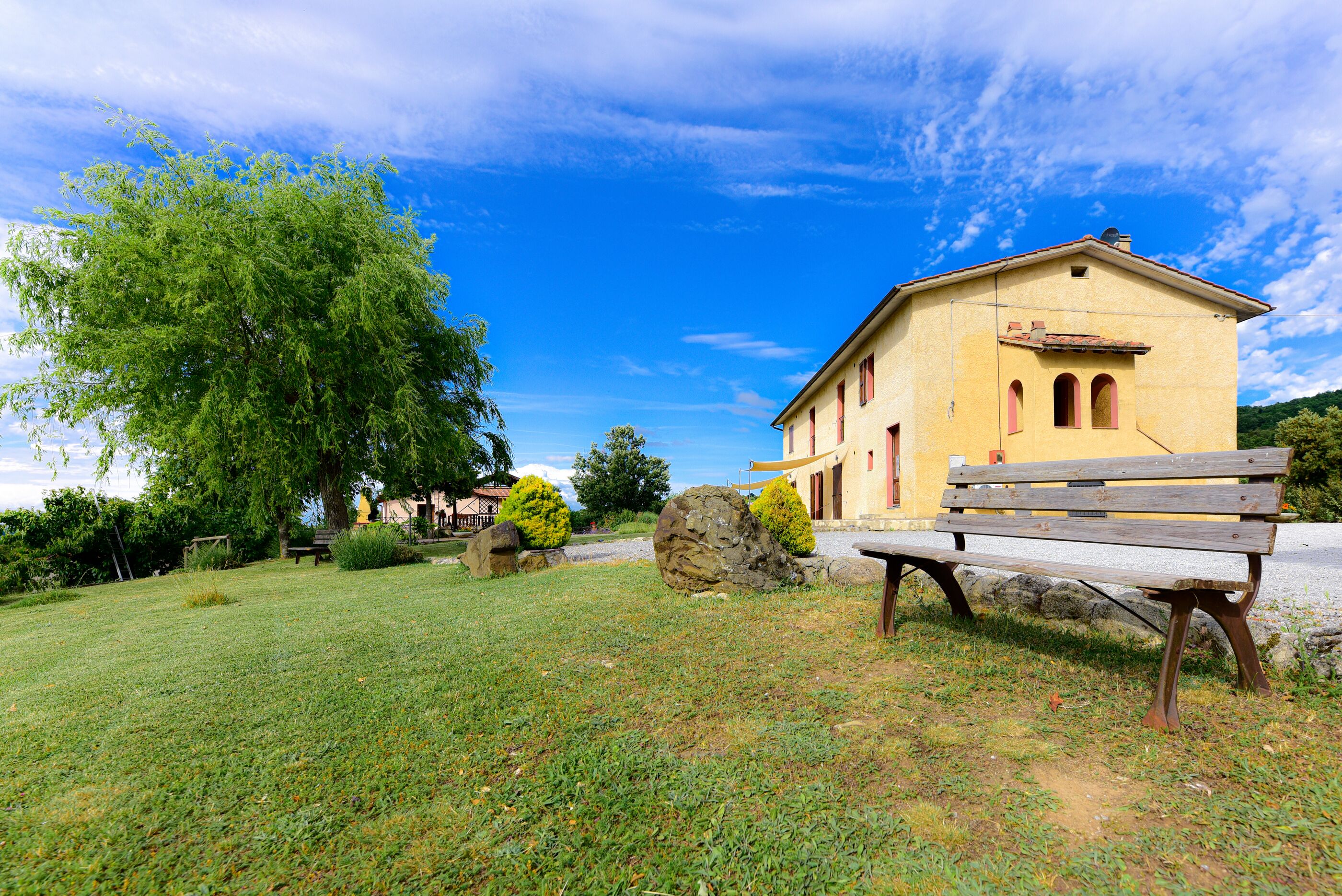 A yellow building with a red roof, a bench, and a large tree in a grassy area.