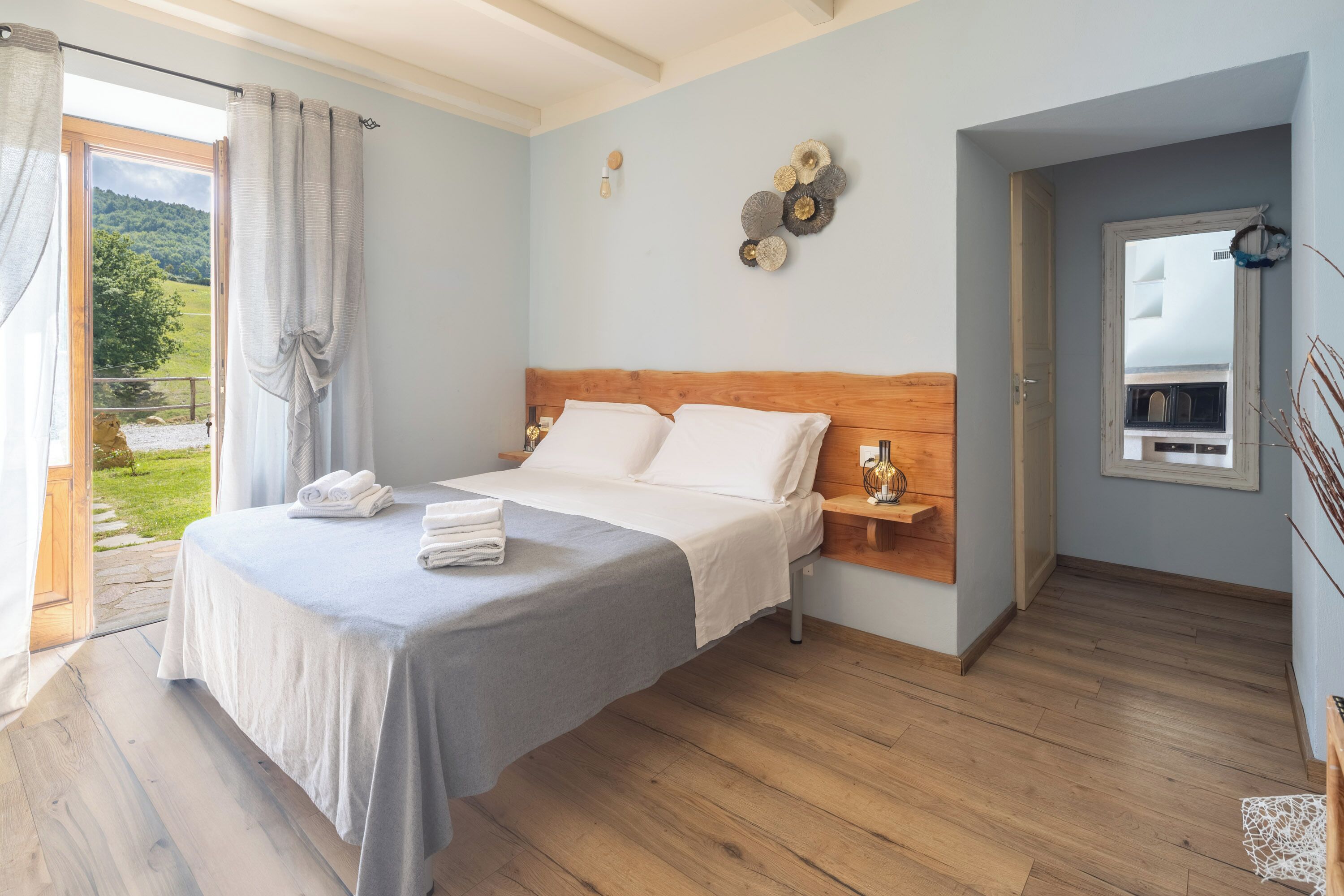 A bedroom with a wooden bed, a bedside table, a view of greenery outside, and a wall-mounted decoration.
