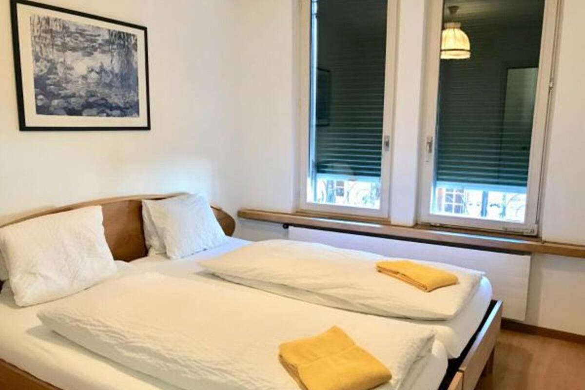 A double bed with white linens and two pillows, a framed picture on the wall, and a window with blinds.