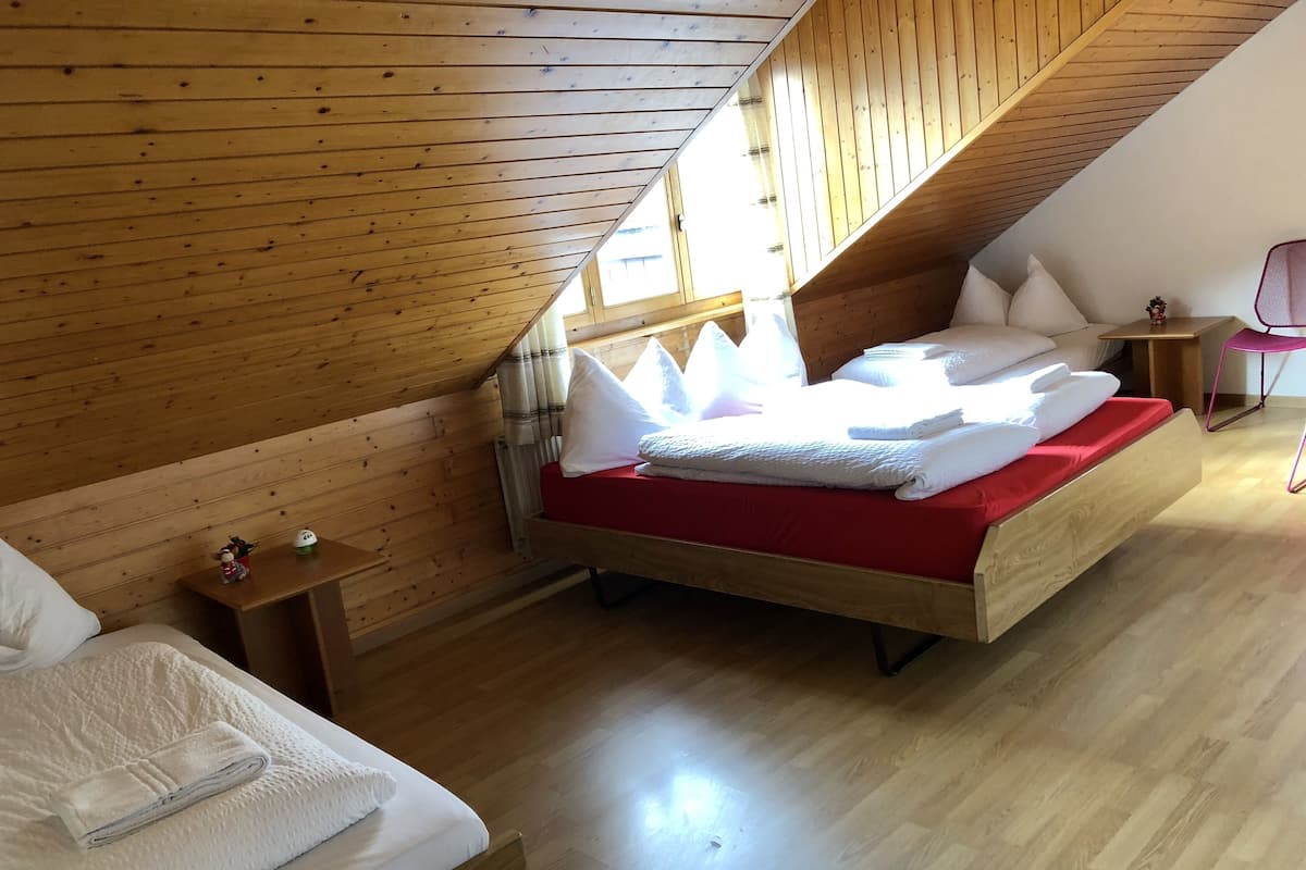 A bedroom with a sloped ceiling, wooden walls, a bed with white linens, a red mattress, and a small bedside table with a lamp.