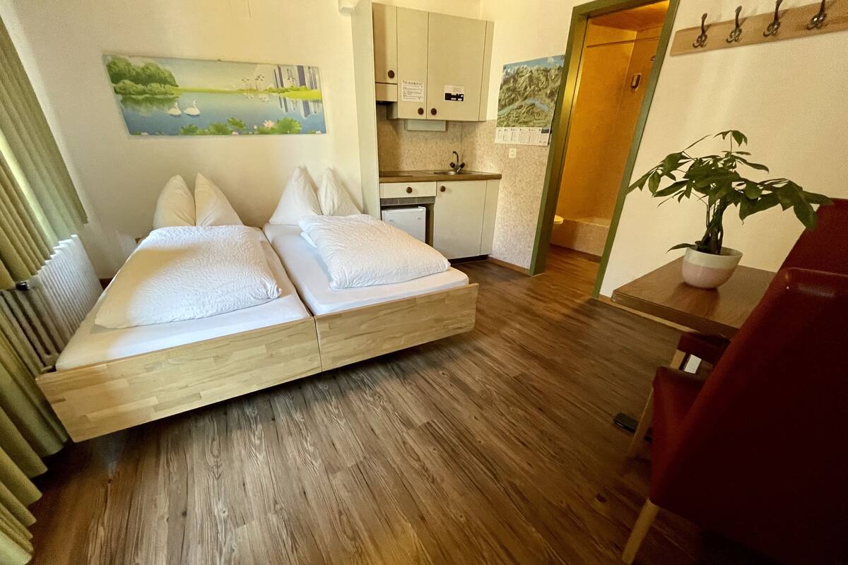A compact room with a wooden floor, a bed with white linens, a small kitchen area, and a potted plant.
