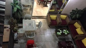 A hotel lobby with a central seating area featuring red chairs and a table with a coffee machine. There are wicker sofas with yellow cushions and a large green plant in the corner. - Hotel Residence Vera (Abidjan)