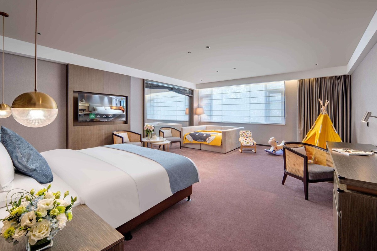 A modern hotel room with a bed, sofa, TV, and a small dining area.