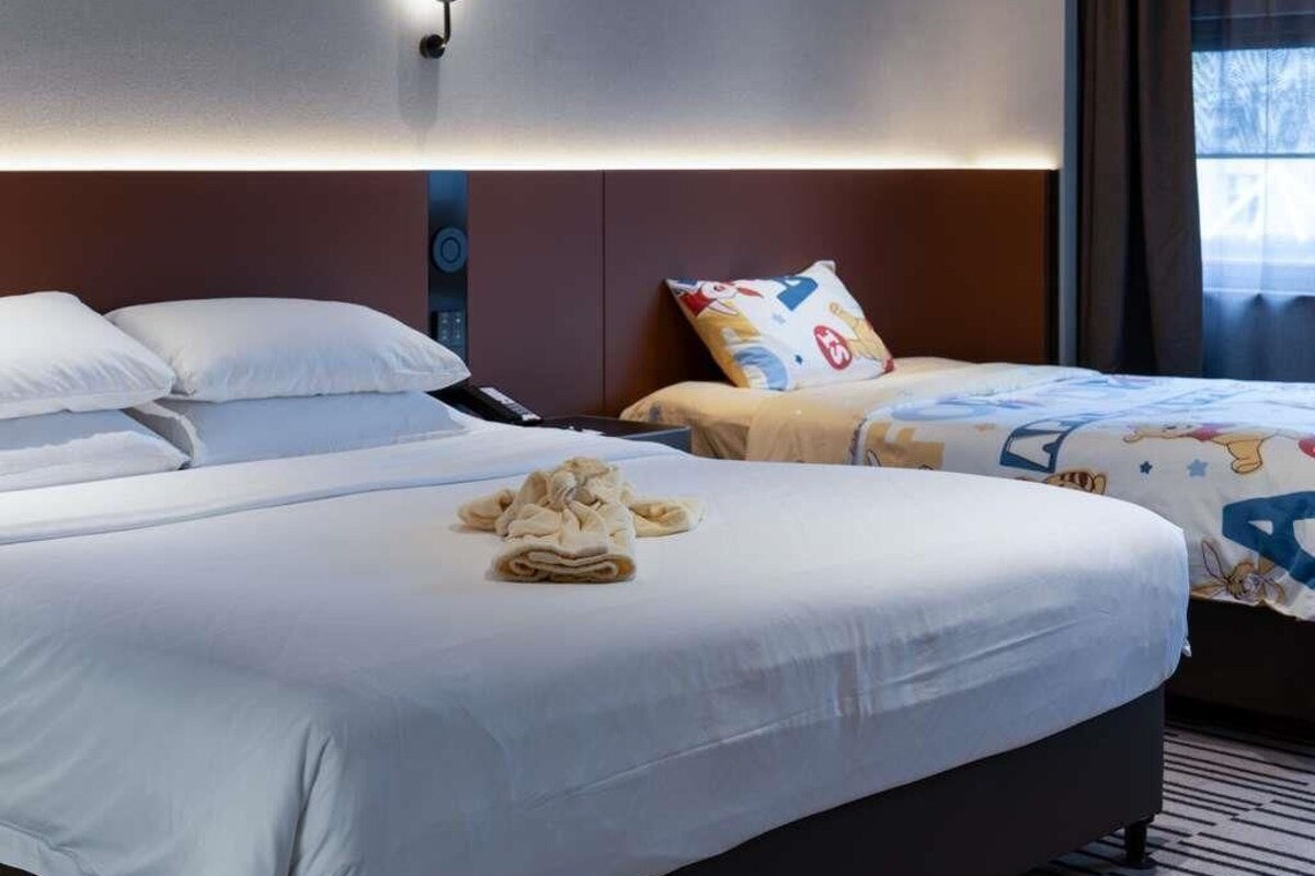 A hotel room with two beds, a bedside table, a wall-mounted light, and a window with curtains.