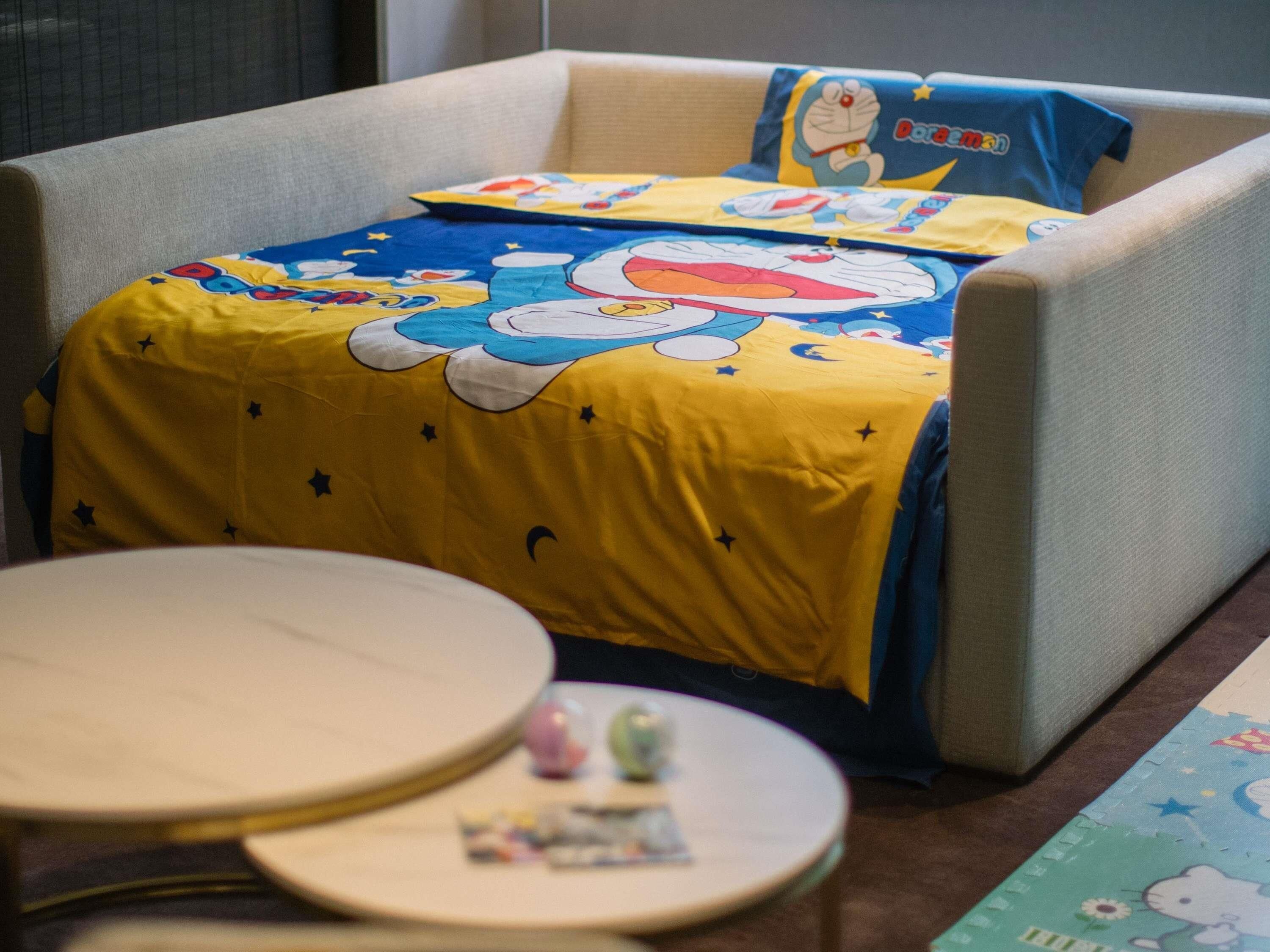 A room with a bed featuring a Doraemon-themed bedspread and pillow. There is a small round table with a white surface and a few items on it.