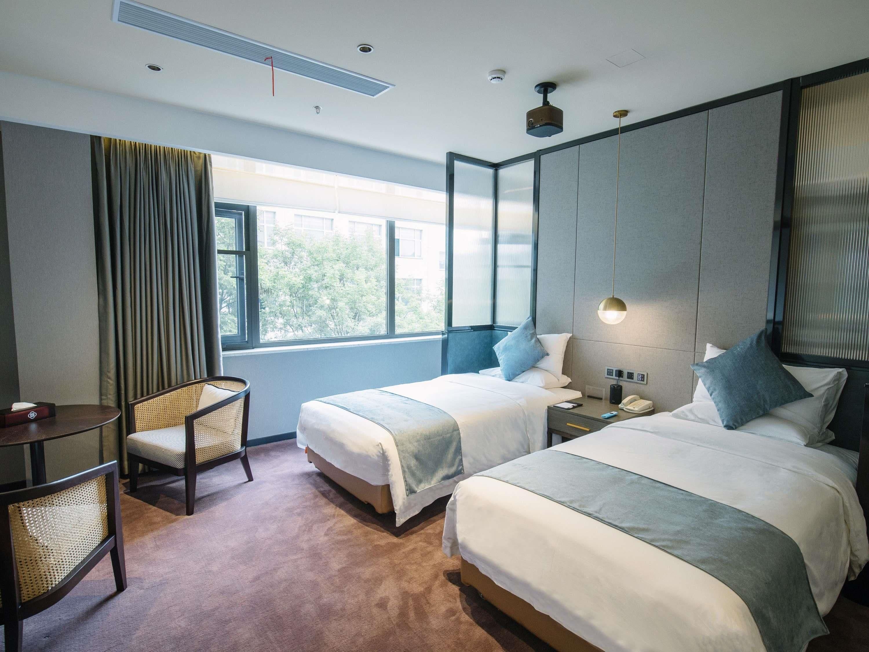 A hotel room with two beds, a small table, a chair, a bedside table with a lamp, and a window with curtains.
