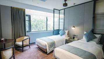 A hotel room with two beds, a small table, a chair, a bedside table with a lamp, and a window with curtains.