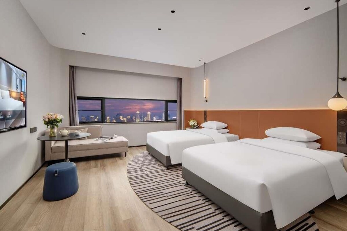 A modern hotel room with two beds, a seating area, a large window with a city view, and a small table with a vase of flowers.