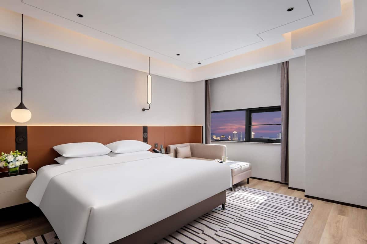 Ein modernes Hotelzimmer mit einem großen Bett, Nachttischen, einem Sofa, einem Fenster mit Blick auf die Stadt und einem gemusterten Teppich.