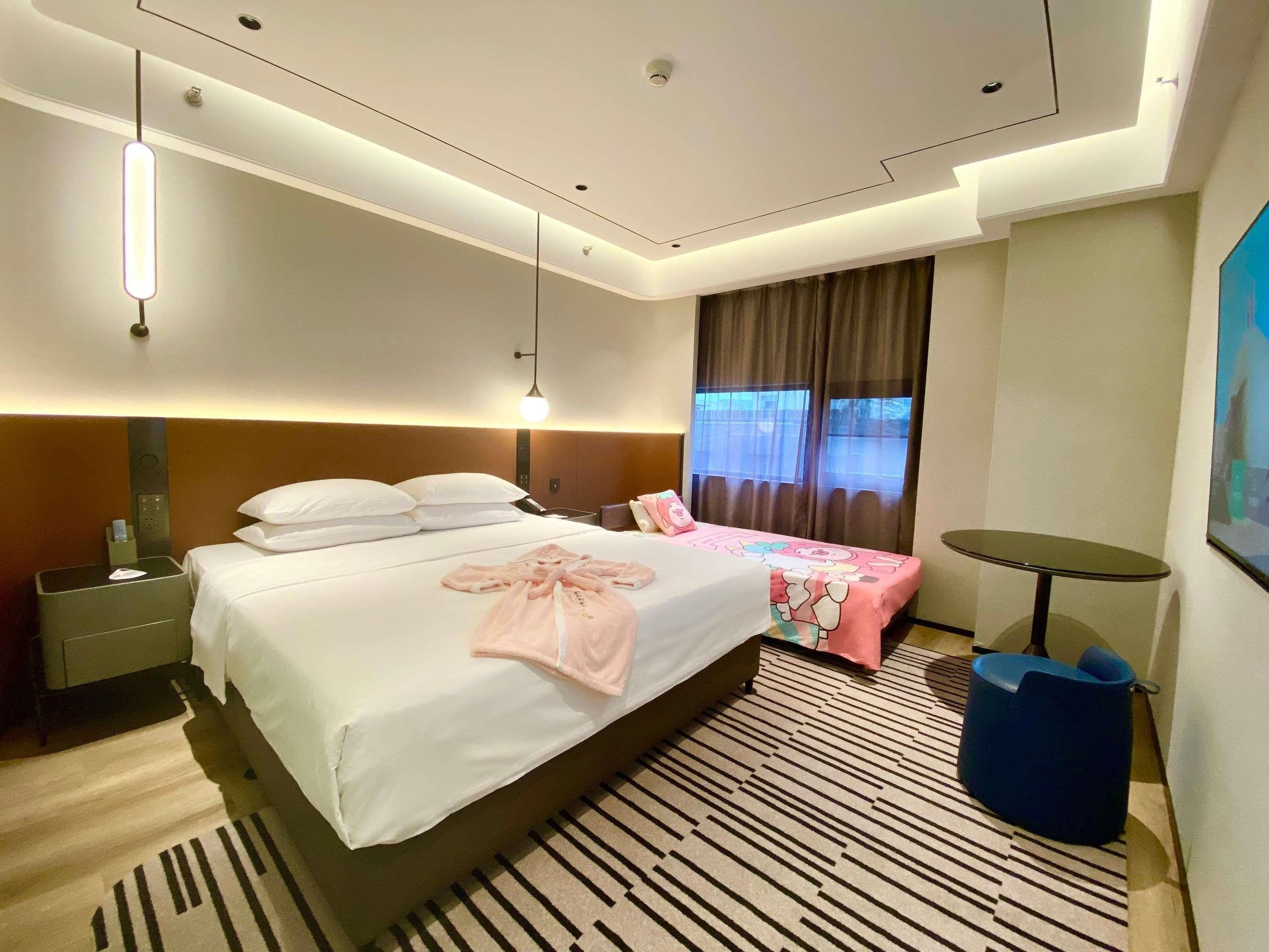 A hotel room with a large bed, bedside tables, a desk, a chair, a small round table, and a window with curtains.