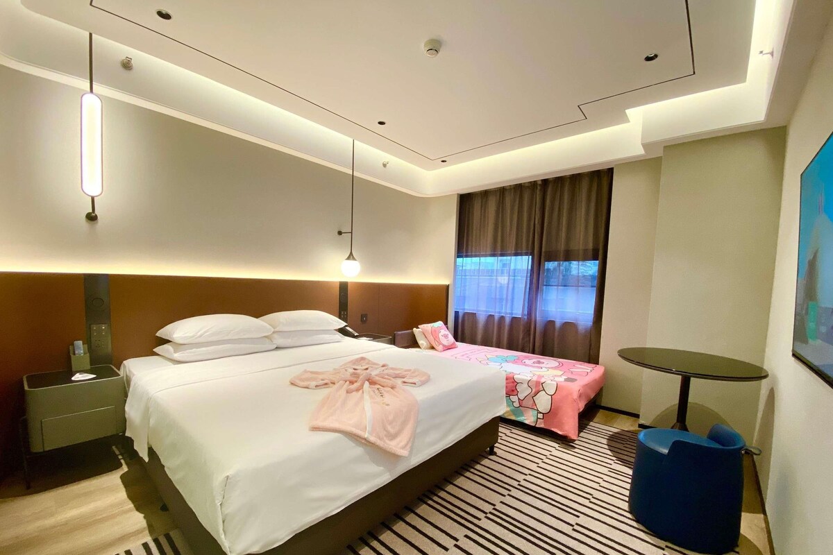 A hotel room with a large bed, bedside tables, a desk, a chair, a small round table, and a window with curtains.