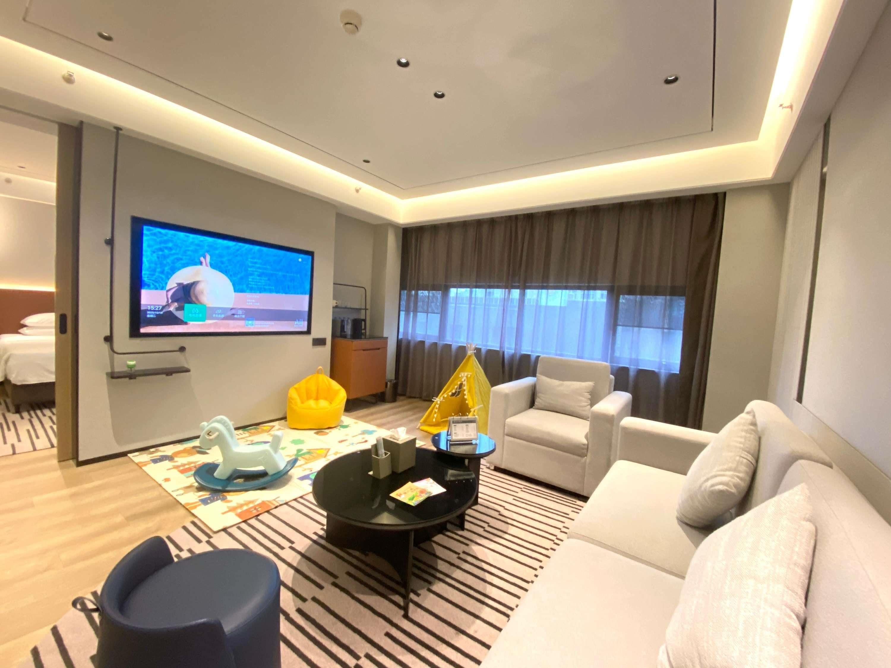 A modern hotel room with a large flat-screen TV, a sofa, a chair, a small table, and a window with curtains.