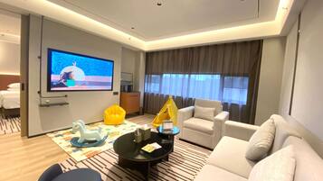 A modern hotel room with a large flat-screen TV, a sofa, a chair, a small table, and a window with curtains.