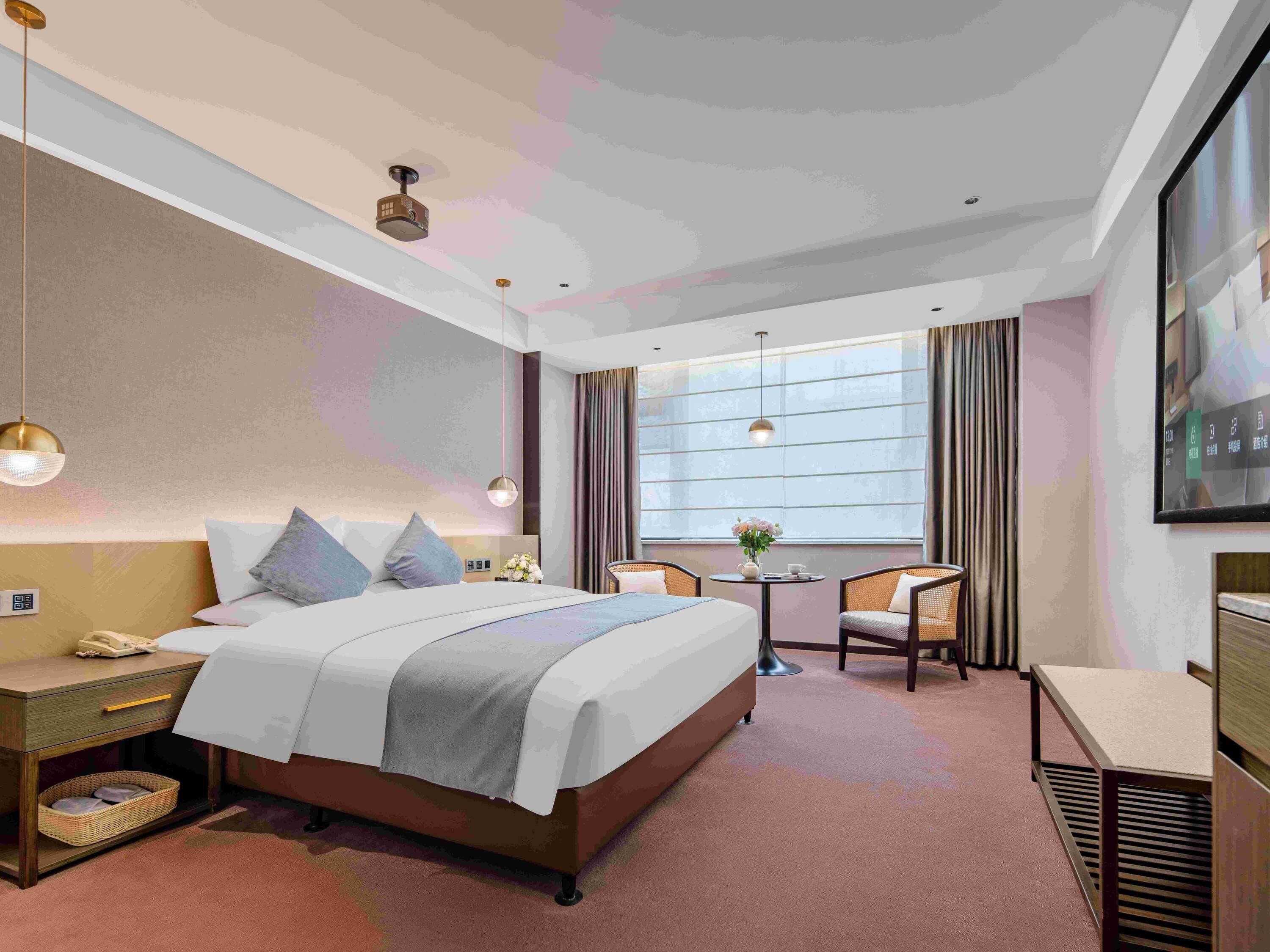 A hotel room with a bed, bedside table, lamp, chair, and a small table with a vase.