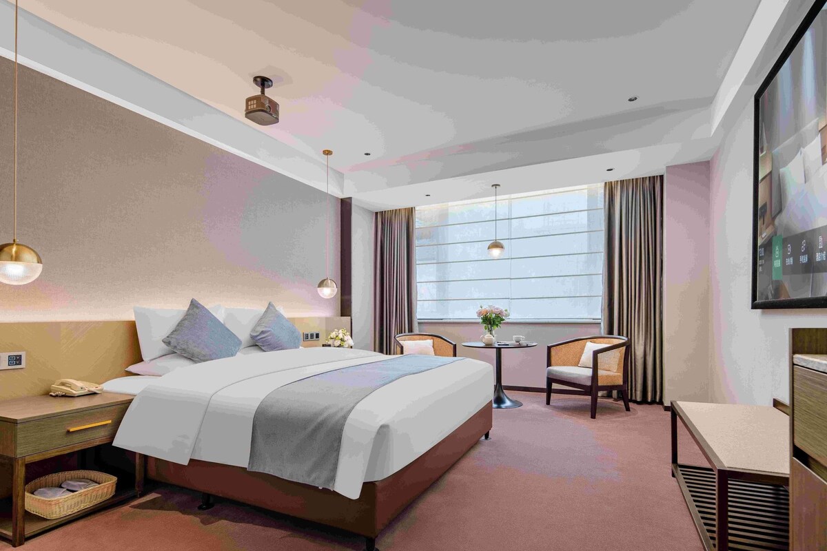 A hotel room with a bed, bedside table, lamp, chair, and a small table with a vase.