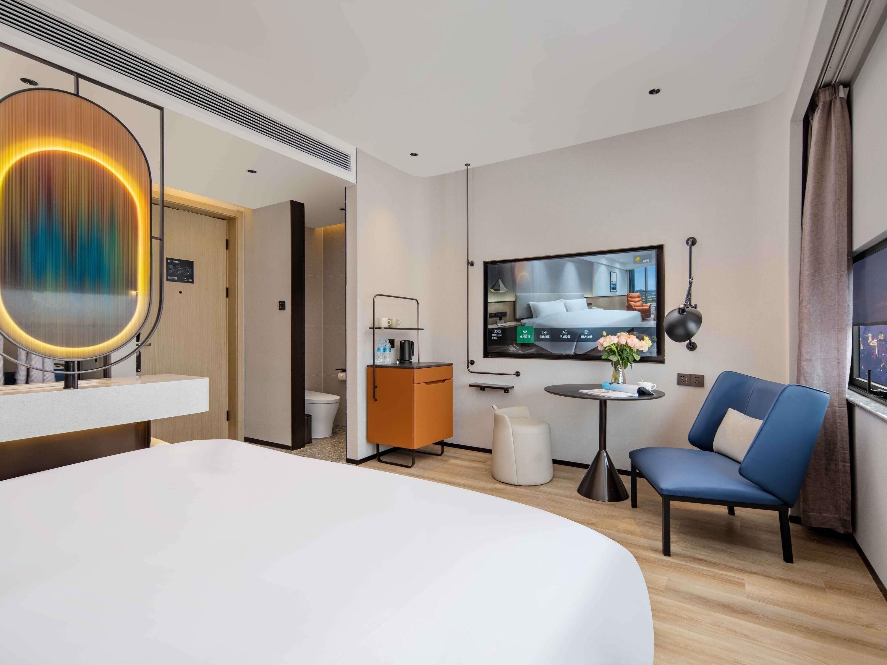 A modern hotel room with a large bed, a small table with a vase, a blue chair, a TV, and a bathroom visible through an open door.