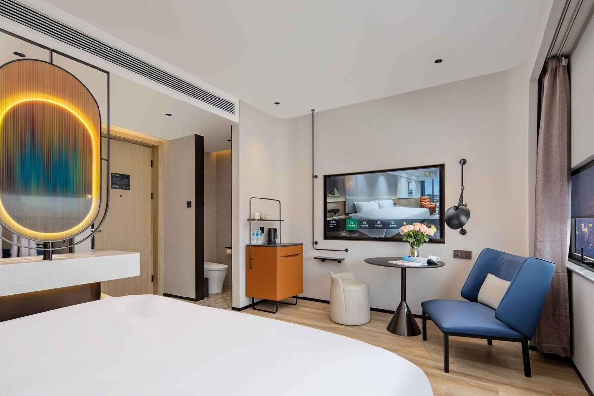 A modern hotel room with a large bed, a small table with a vase, a blue chair, a TV, and a bathroom visible through an open door.