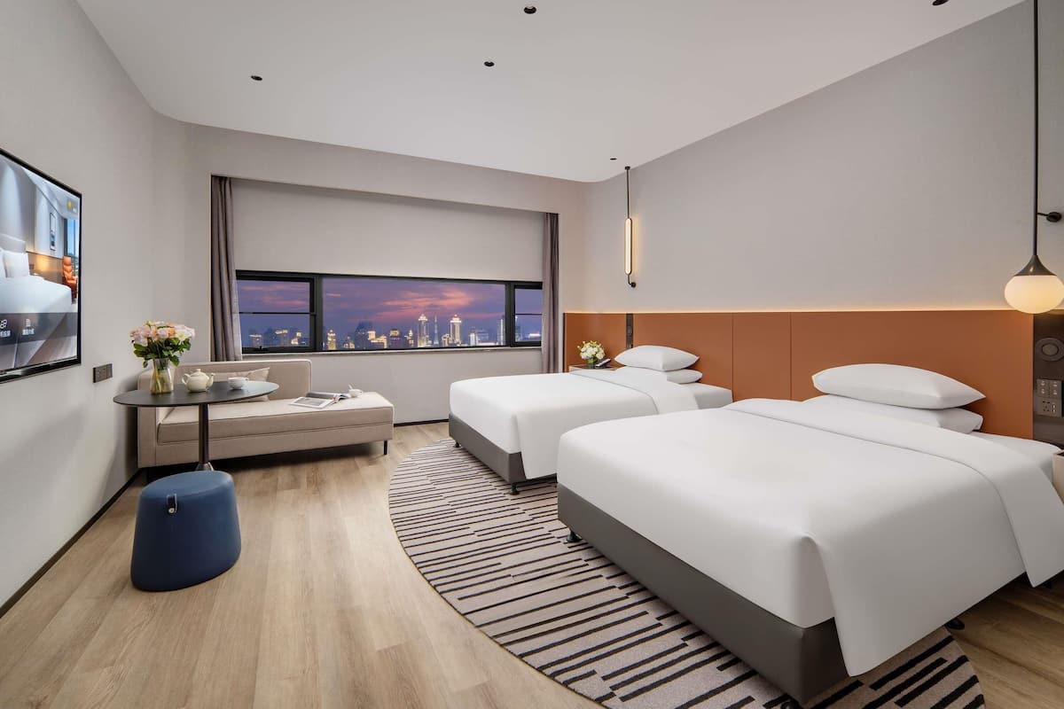 Ein modernes Hotelzimmer mit Blick auf die Stadt, inklusive einer Couch, zwei Betten, einem runden Tisch und einem blauen Ottomanen.