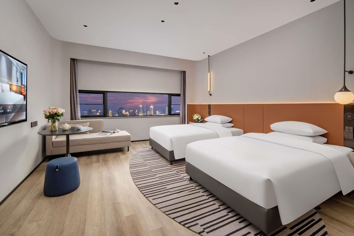 A modern hotel room with a city view, a sofa, two beds, a round table, and a blue ottoman.
