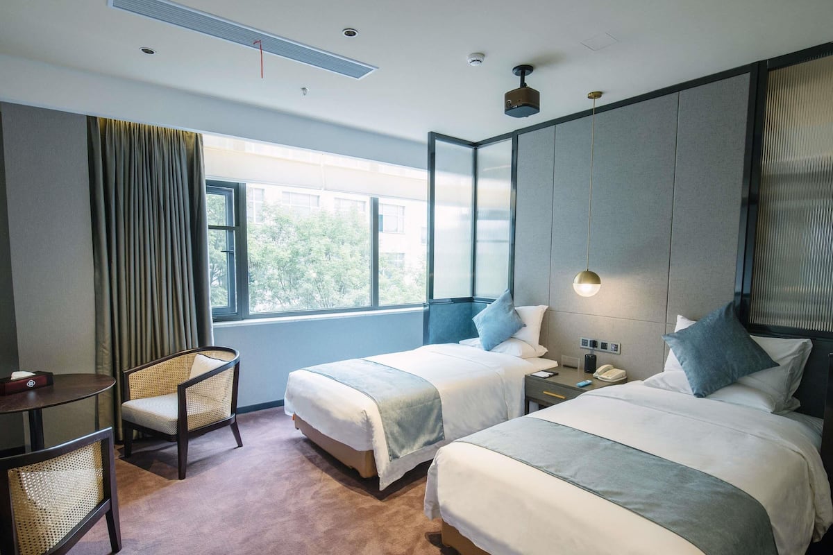Ein Hotelzimmer mit zwei Betten, einem kleinen Tisch, einem Stuhl, einem Nachttisch mit Lampe und einem fenster mit Vorhängen.
