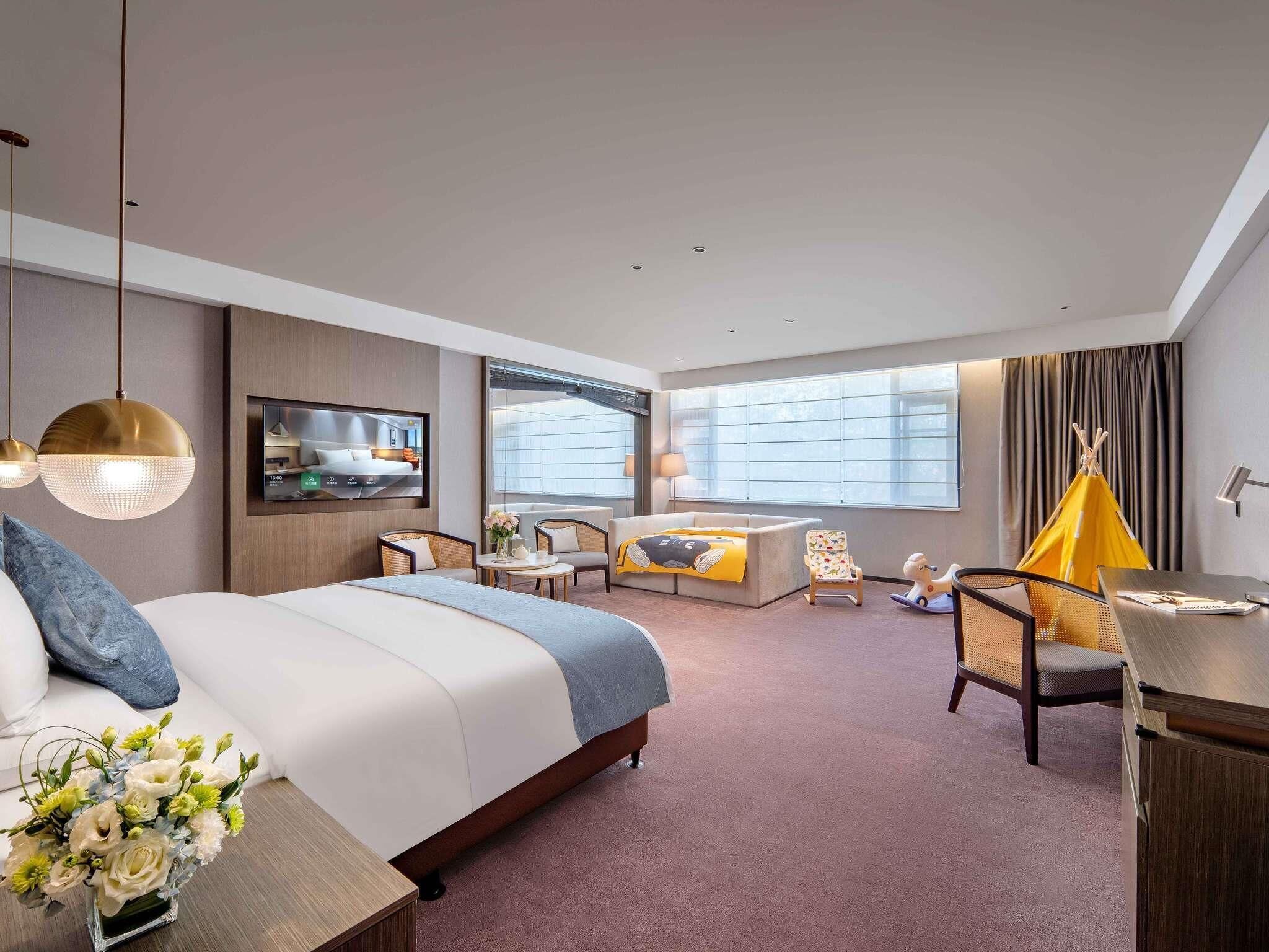 A modern hotel room with a large bed, a TV, a sofa, a desk, and a chair.