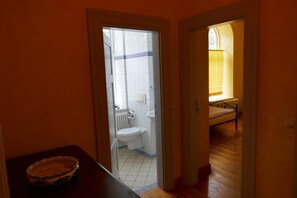Een smalle gang die leidt naar een badkamer met toilet, wastafel en een bed.