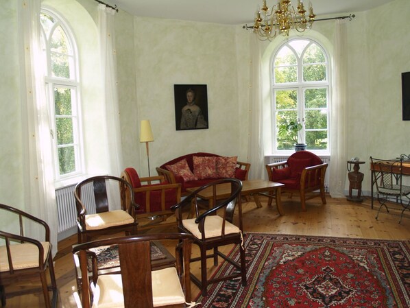 Ein Zimmer mit einem roten Sofa, Holzstühlen, einem Tisch und einem großen Teppich. Zwei Fenster mit Vorhängen, eine Kristallleuchter und ein gerahmtes Bild sind an den Wänden zu sehen.