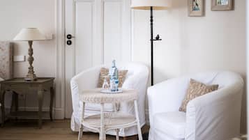 Une pièce avec une porte blanche, une table en osier sur laquelle se trouvent une bouteille et des verres, un fauteuil blanc, un lampadaire et une petite table avec une lampe.