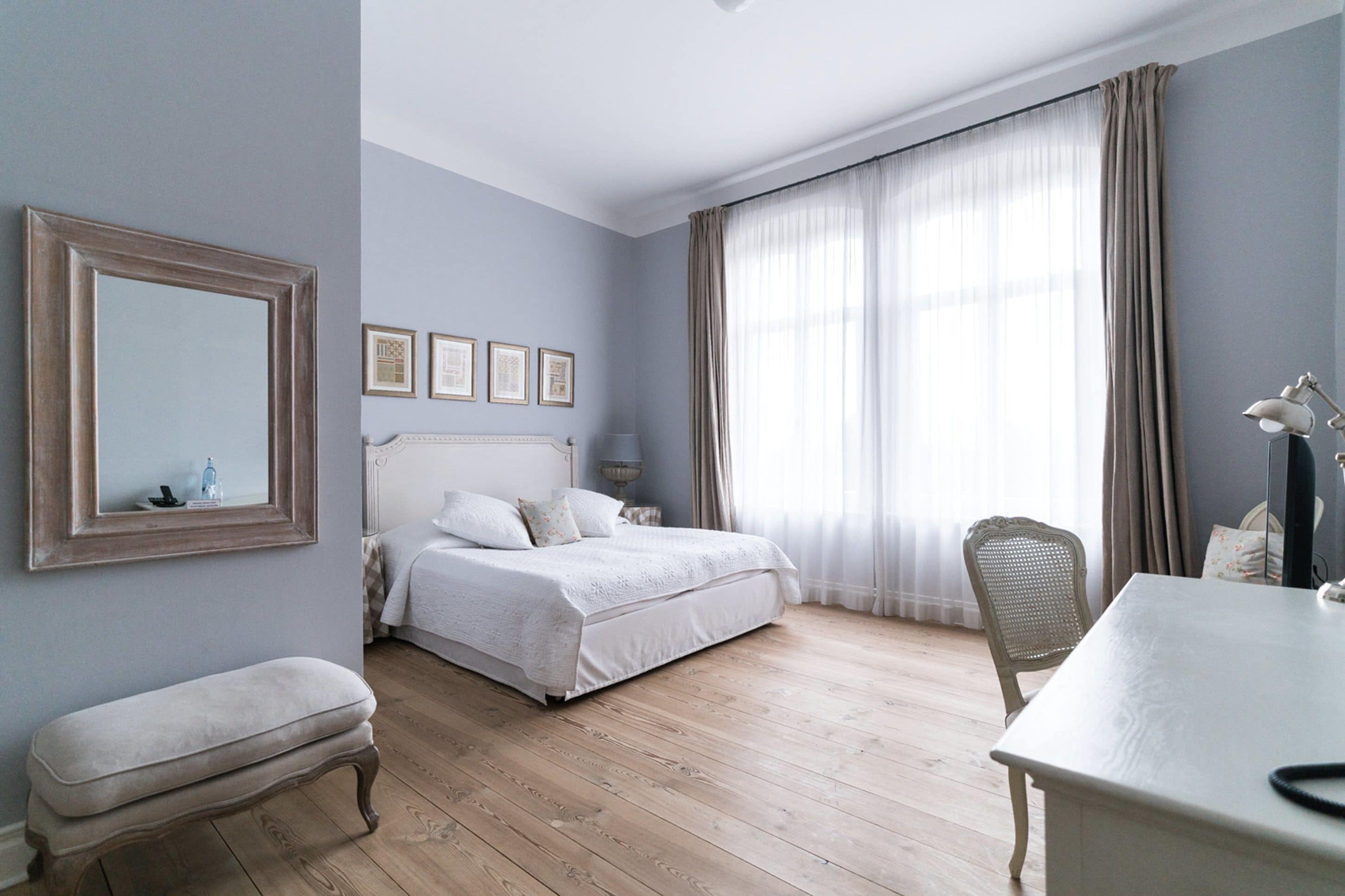 Ein Schlafzimmer mit Bett, Nachttisch, Sessel, Schreibtisch, Spiegel und gerahmten Bildern an der Wand.