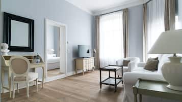 Habitación de hotel con piso de madera, un escritorio blanco con una silla, televisor, sofá y una lámpara.