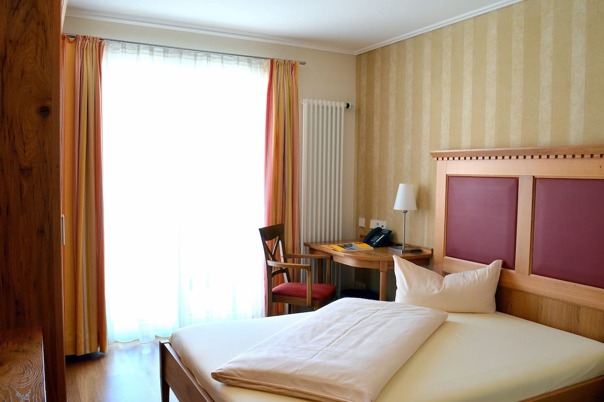 A hotel room with a bed, a desk with a chair, a telephone, and a window with curtains.