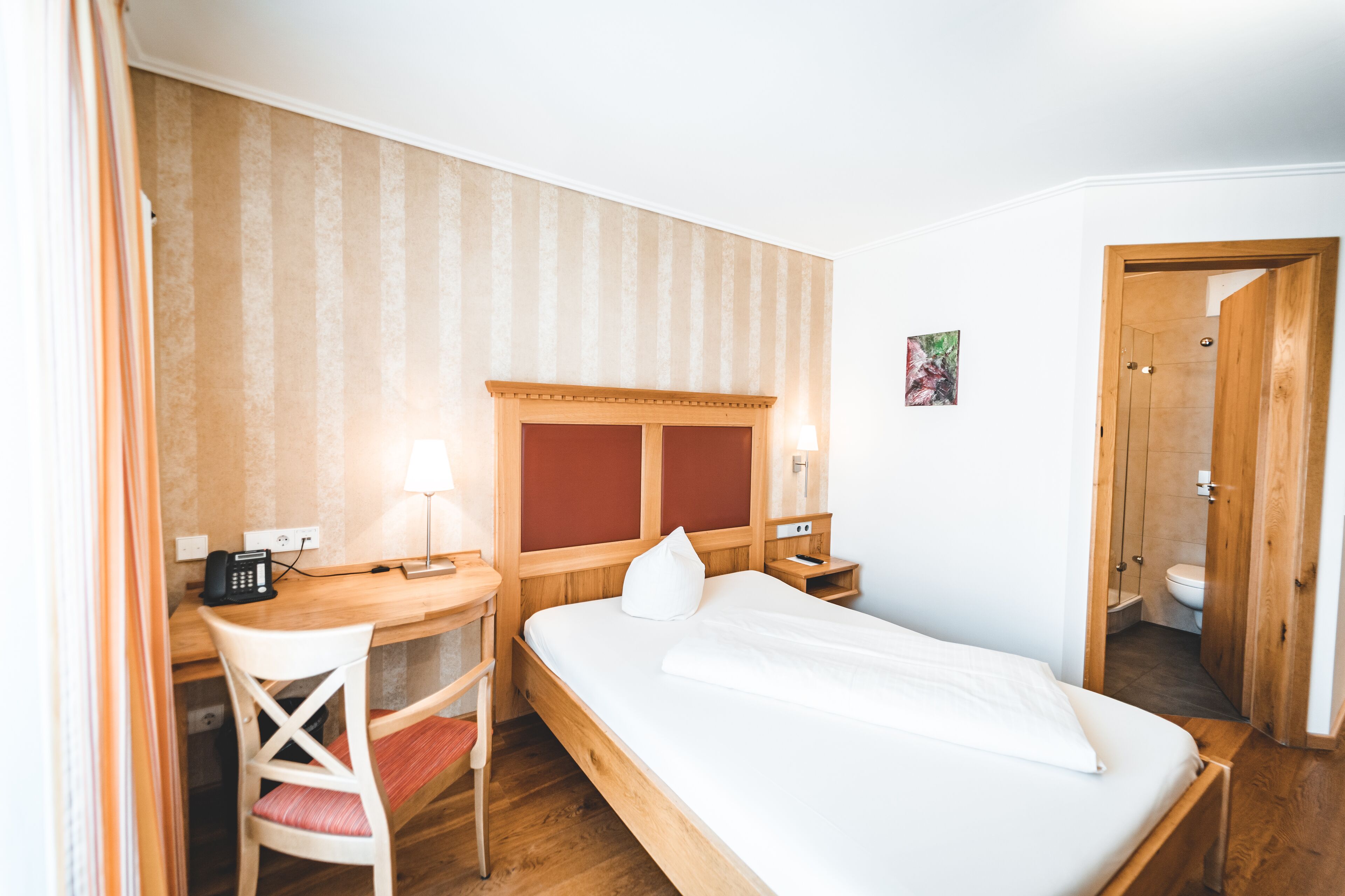 A hotel room with a wooden bed, a desk with a chair, a telephone, and a nightstand with a lamp.