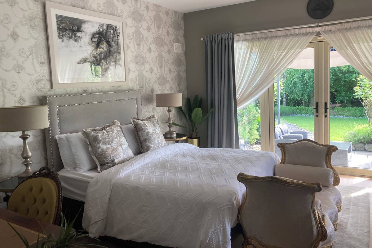A bedroom with a large bed, two bedside tables with lamps, a chair, a framed artwork on the wall, and a view of the garden through the window.