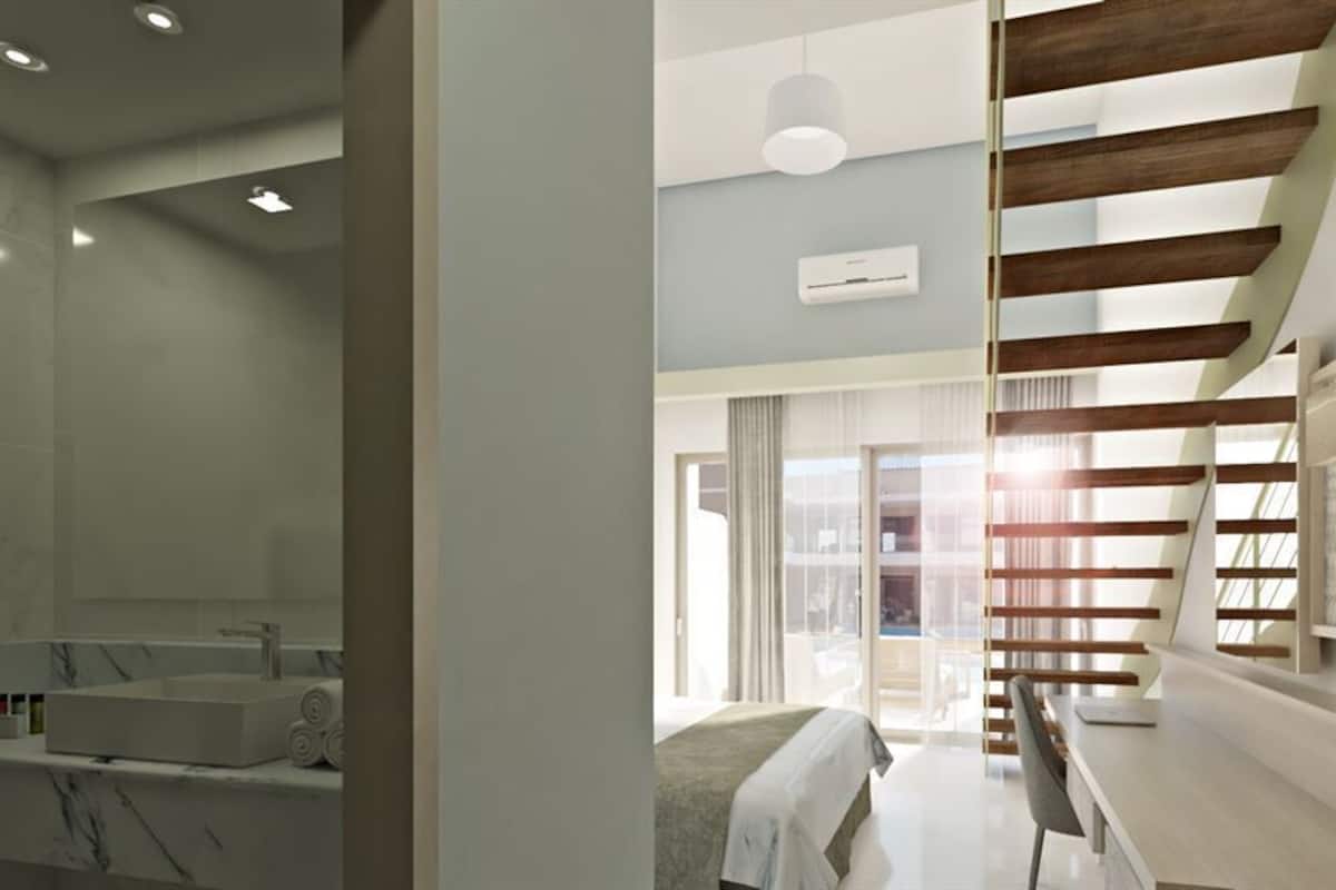 A modern hotel room with a marble bathroom, a bed with a view, and a staircase with wooden beams.