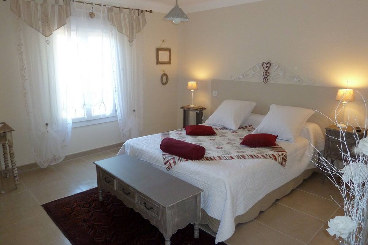 Un dormitorio con una cama grande, dos mesitas de noche, un tocador, una ventana con cortinas y una lámpara colgante.