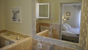 Un bany amb un mirall gran, un lavabo i un vàter. Hi ha un quadre al mur i es veu un llit a través del mirall.