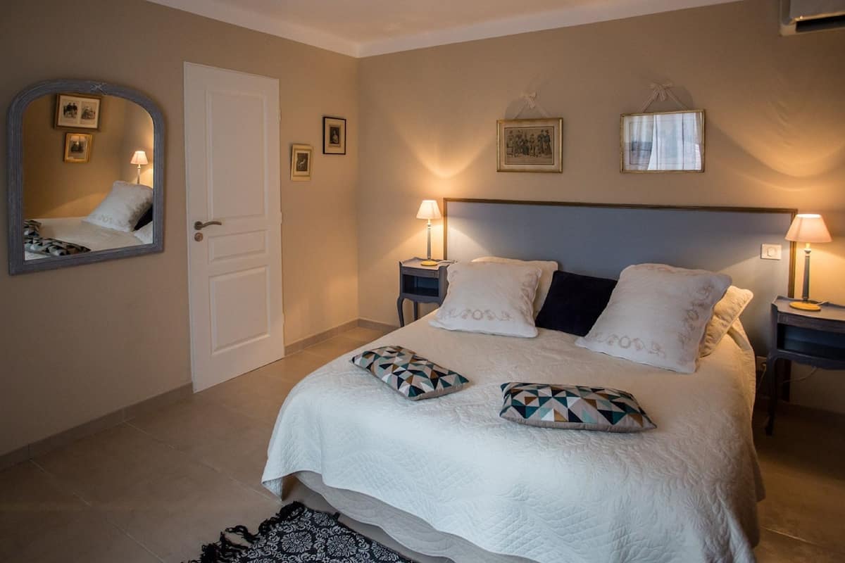 Un dormitorio ordenado con una cama, dos mesitas de noche con lámparas, un espejo y cuadros enmarcados en la pared.