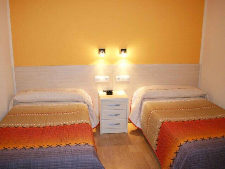 A room with two single beds, a small white nightstand with drawers, and two wall-mounted light fixtures.