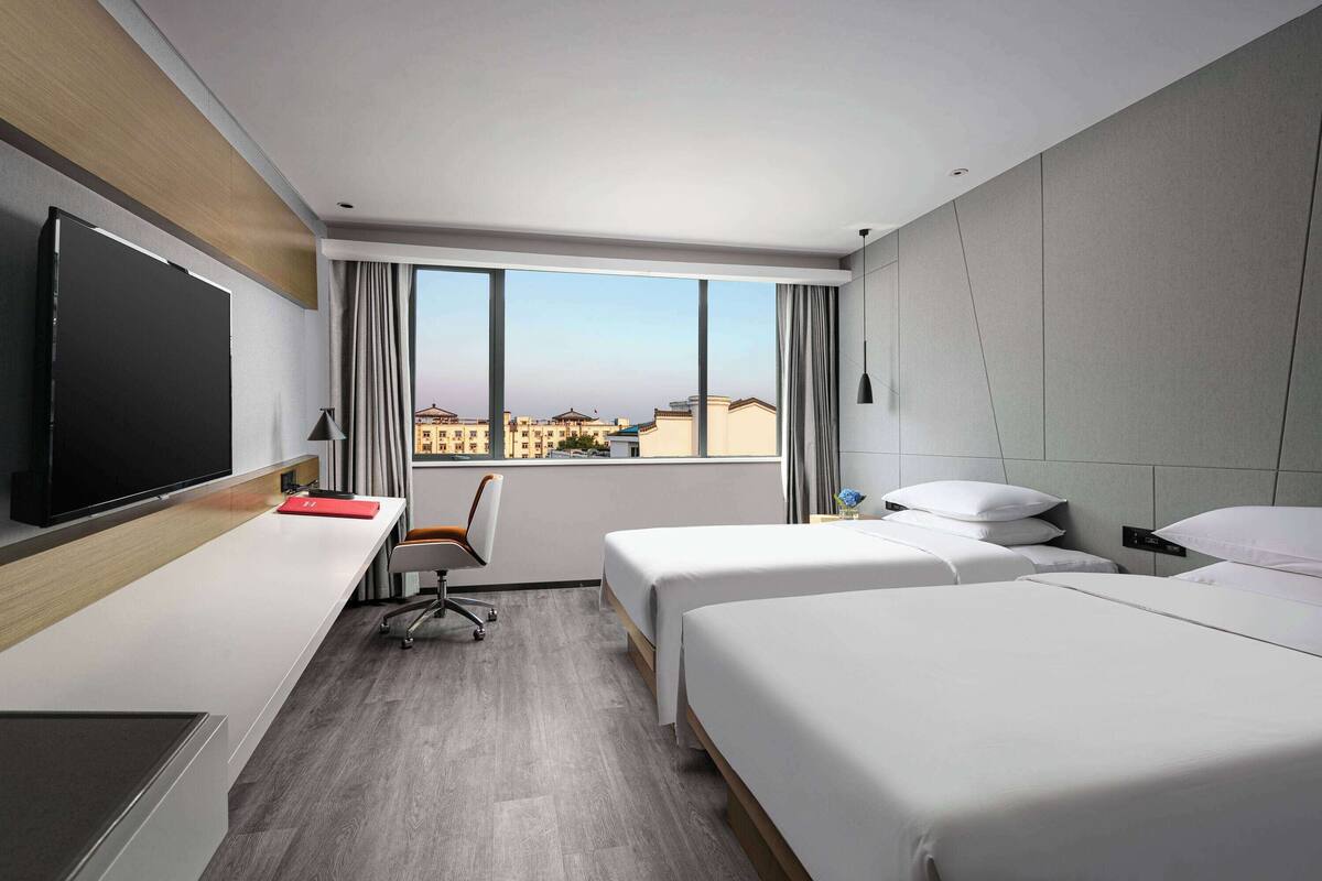 Ein modernes Hotelzimmer mit großem Fernseher, Schreibtisch, zwei Betten und Blick auf Gebäude durch große Fenster.