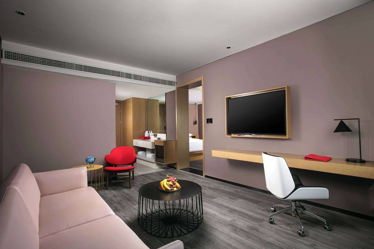 Ein modernes Hotelzimmer mit einer Couch, einem runden Couchtisch mit Obst, einem roten Sessel, einem Schreibtisch mit einem Computer, einem Fernseher und einem Spiegel.