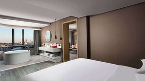 Een moderne hotelkamer met een groot bed, een aparte badkamer met badkuip, een wastafel met spiegel en uitzicht op de stad door grote ramen.