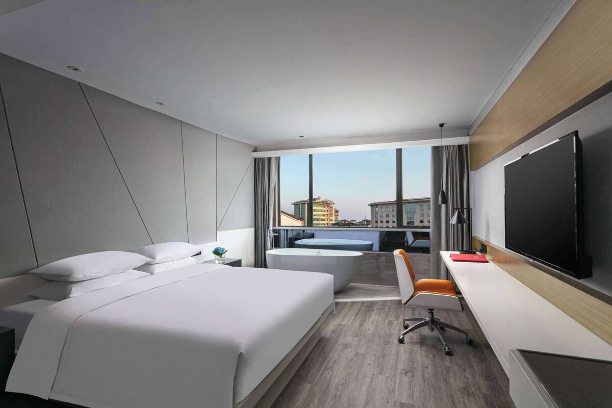 Ein modernes Hotelzimmer mit einem großen Bett, einem Schreibtisch, einem Sessel, einem Flachbildfernseher und Blick auf die Stadt.