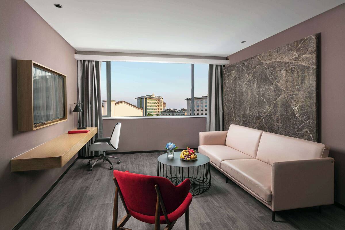 Ein modernes Hotelzimmer mit großem Fenster, einer Couch, einem roten Sessel, einem Schreibtisch und einem runden Couchtisch.