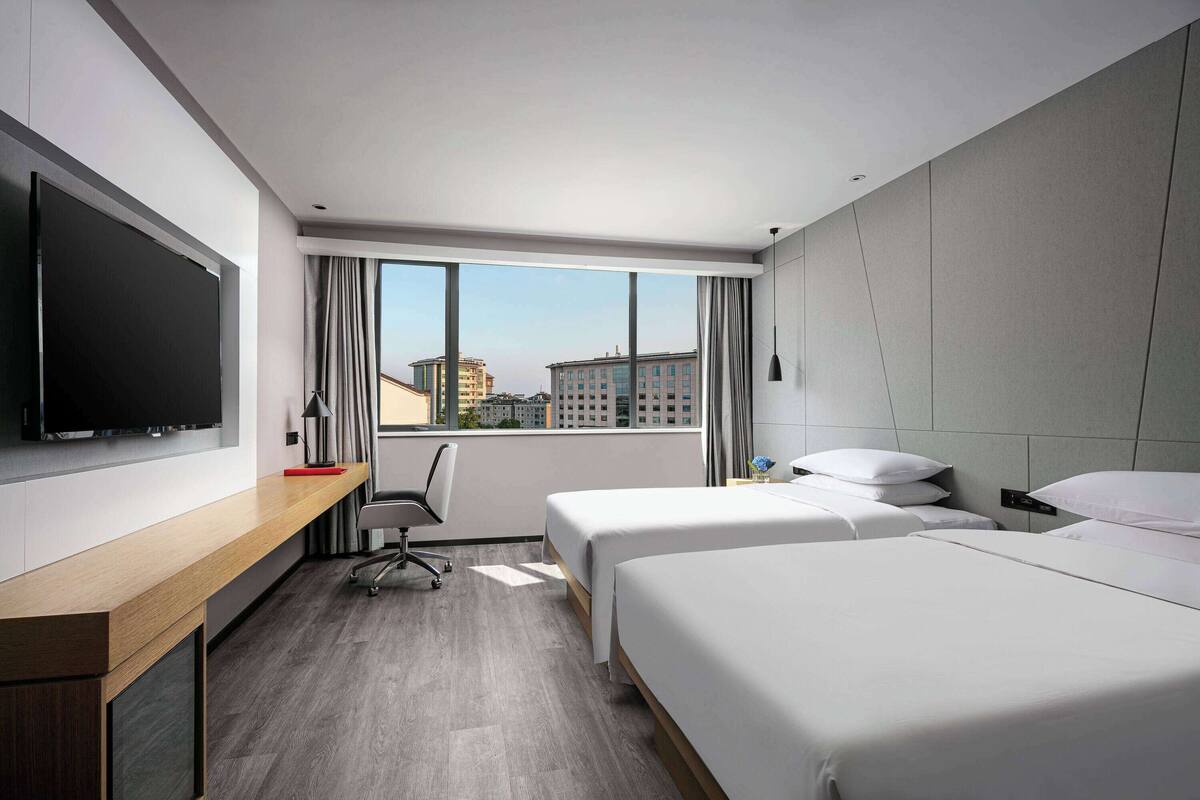 Ein Hotelzimmer mit großem Fenster, einem Schreibtisch mit Flachbildfernseher, zwei Betten und einem Sessel.