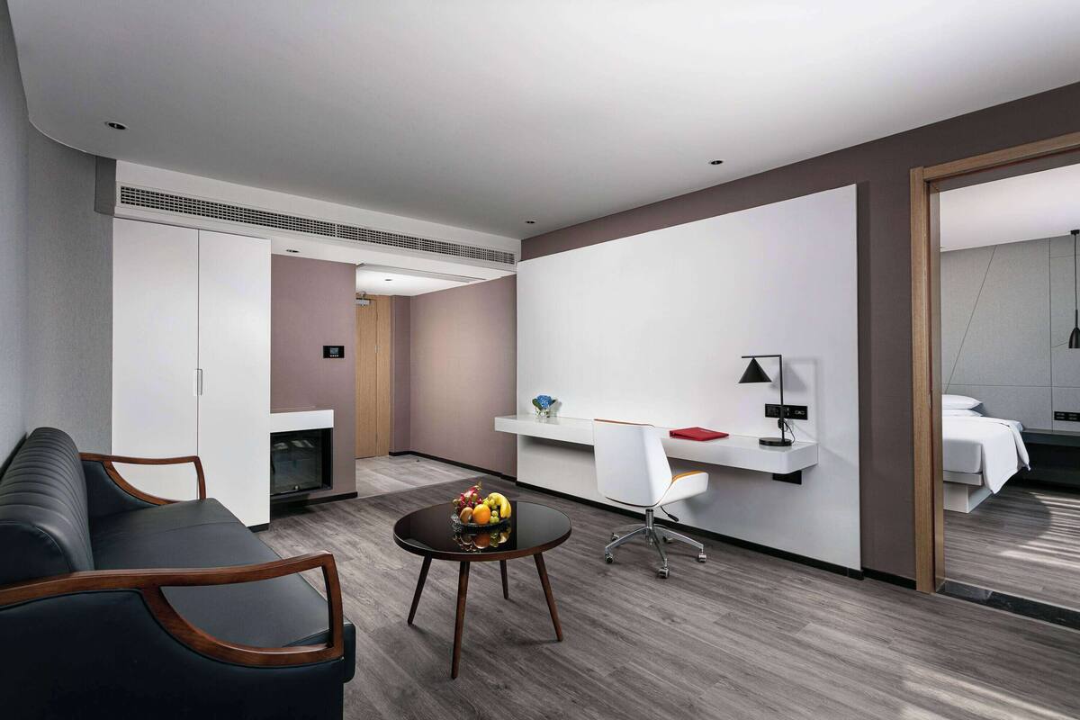 Ein modernes Hotelzimmer mit einer Couch, einem runden Tisch mit Obst, einem Schreibtisch mit Stuhl und einem Bett.