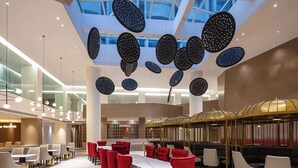 Een ruime hotellobby met een hoge plafond, moderne kroonluchters en een centraal zitgedeelte met rode en beige stoelen.