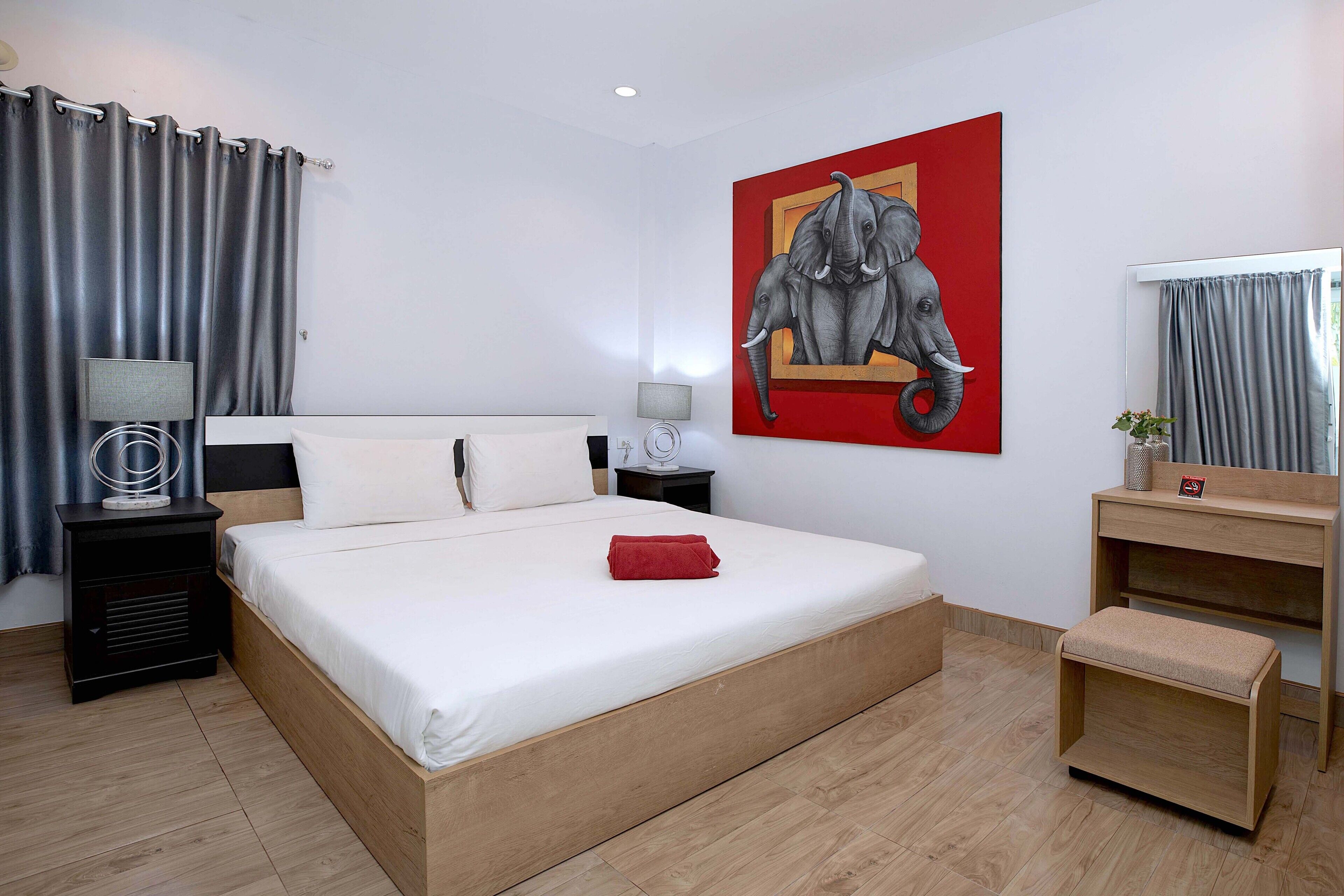 Ein Hotelzimmer mit einem großen Bett, einem Nachttisch mit Lampe, einer kleinen Holzbank, einem Spiegel und einem Elefantenbild.